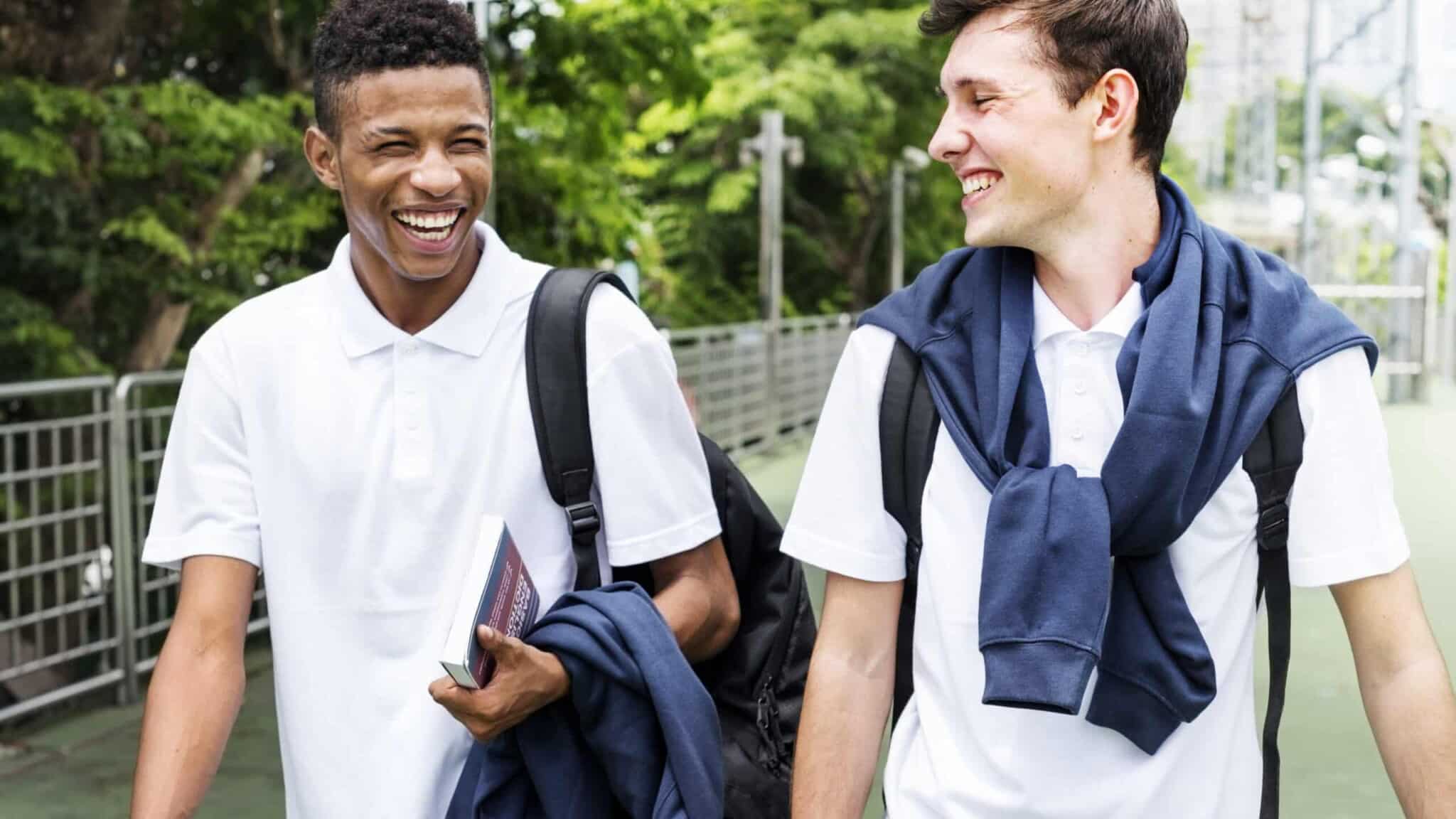 Two smiling students in white uniforms walking outdoors, holding books and backpacks. Their camaraderie and positive energy reflect the supportive academic environment found at a prep school, preparing them for future educational success.