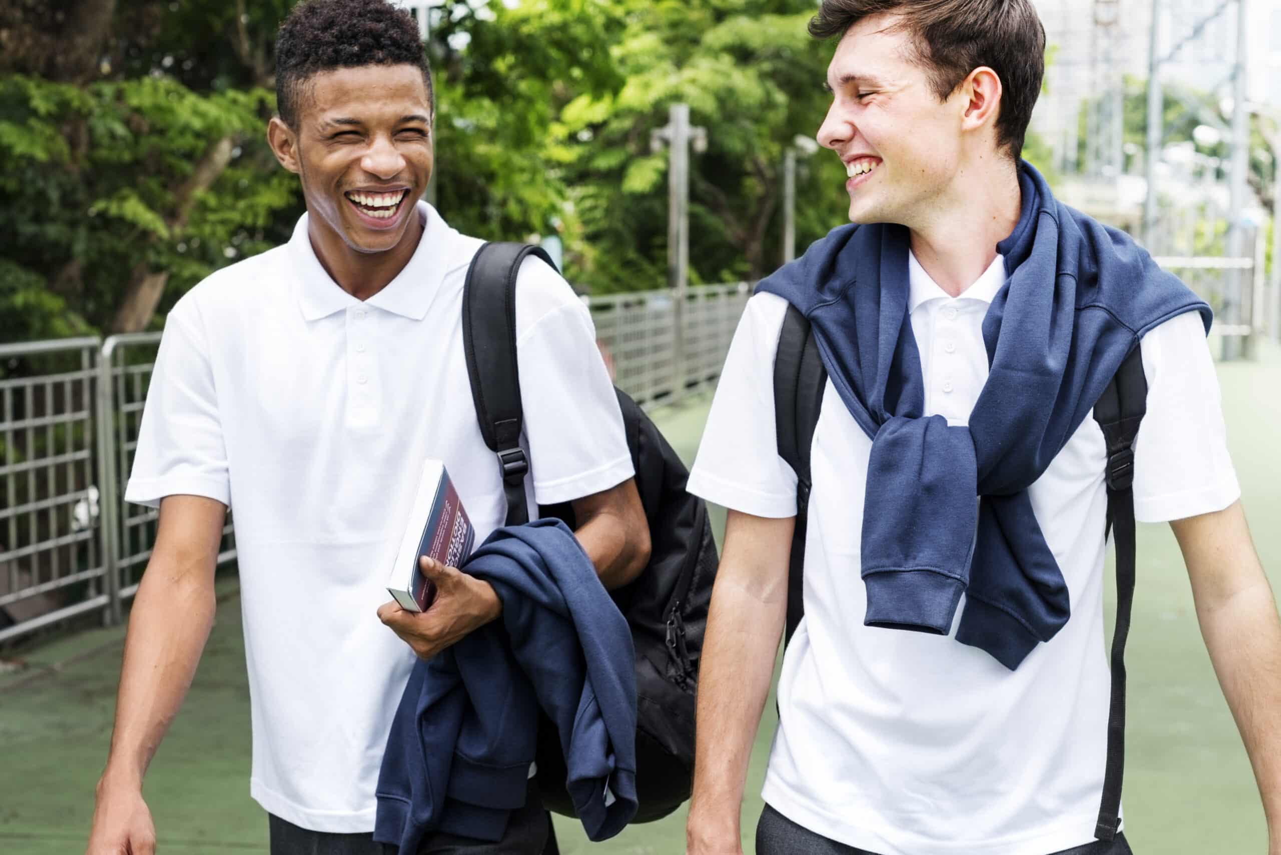 Two smiling students in white uniforms walking outdoors, holding books and backpacks. Their camaraderie and positive energy reflect the supportive academic environment found at a prep school, preparing them for future educational success.