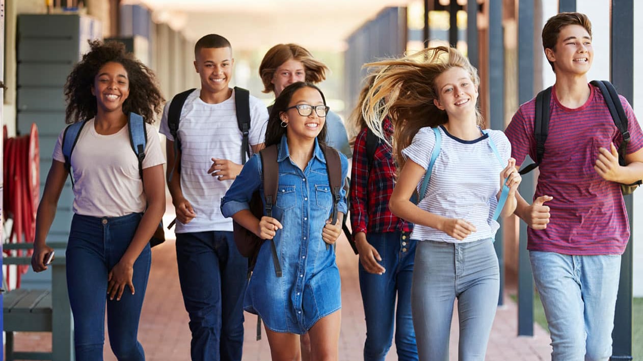 A group of diverse, happy teenagers walking together on a school campus, carrying backpacks, representing enthusiasm and readiness for the future. This image symbolizes excitement for higher education and the importance of early college planning.