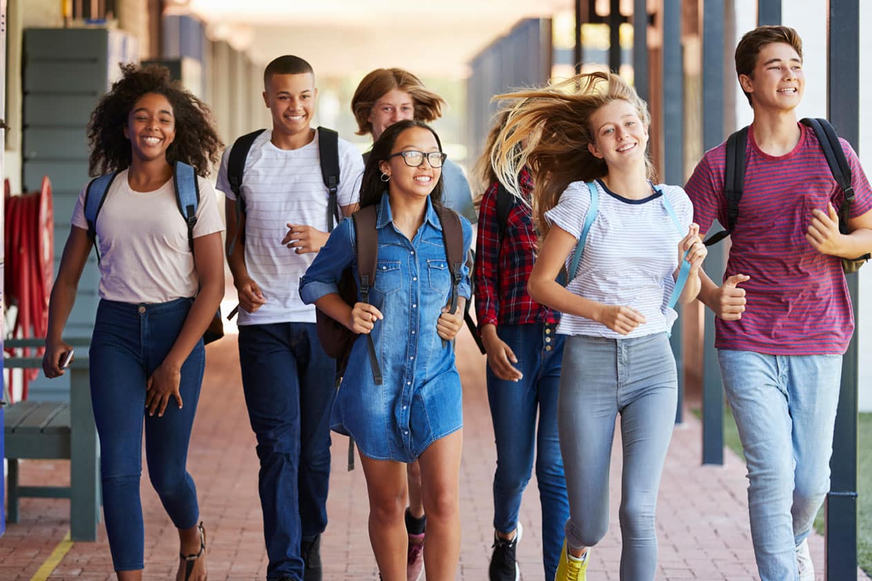 A group of diverse, happy teenagers walking together on a school campus, carrying backpacks, representing enthusiasm and readiness for the future. This image symbolizes excitement for higher education and the importance of early college planning.